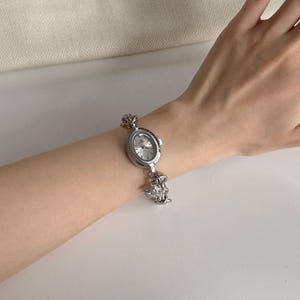 May include: Silver oval watch with a decorative crown design on the band. The watch face is silver with black hands and markers. The watch is worn on a wrist.