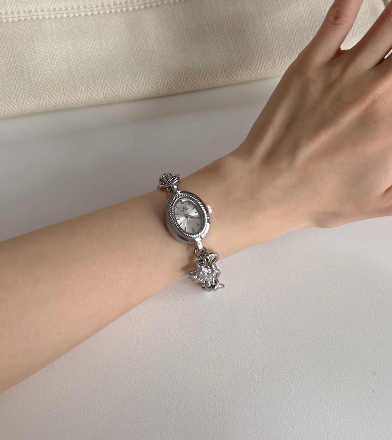 May include: Silver oval watch with a decorative crown design on the band. The watch face is silver with black hands and markers. The watch is worn on a wrist.