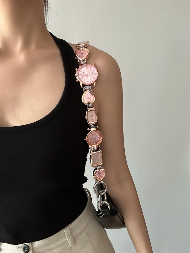 May include: A pink and gold watch strap with multiple watch faces. The strap is made of metal and has a heart-shaped design. The watch faces are all different sizes and shapes.