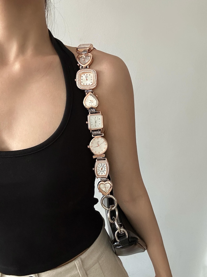 May include: A gold-tone chain bag strap with multiple small watch faces attached. The strap is made up of linked hearts and square watch faces.