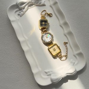 May include: A gold-tone watch bracelet with three square watch faces. The watch faces have different designs, including a black face with gold hands, a white face with gold hands and Roman numerals, and a gold face with a floral design. The bracelet is on a white ceramic tray with a bow design.