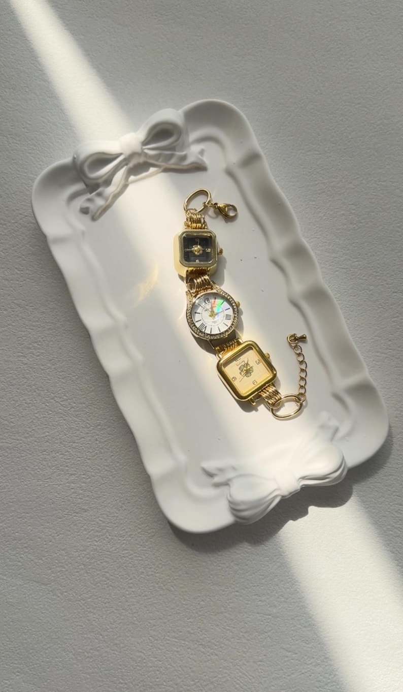 May include: A gold-tone watch bracelet with three square watch faces. The watch faces have different designs, including a black face with gold hands, a white face with gold hands and Roman numerals, and a gold face with a floral design. The bracelet is on a white ceramic tray with a bow design.