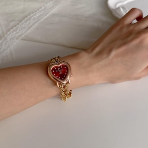 May include: A gold-toned wristwatch with a heart-shaped face and a red dial. The watch features small white dots and a delicate gold chain with a charm. The watch is worn on a wrist.