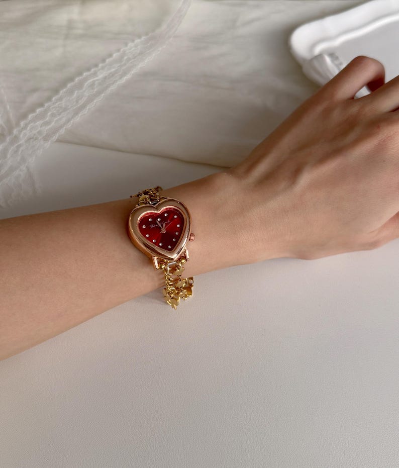 May include: A gold-toned wristwatch with a heart-shaped face and a red dial. The watch features small white dots and a delicate gold chain with a charm. The watch is worn on a wrist.