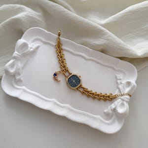 May include: A gold-toned wristwatch with a dark blue face and a decorative moon and star charm. The watch is displayed on a white, bow-shaped tray. The watch band has a unique chain design.