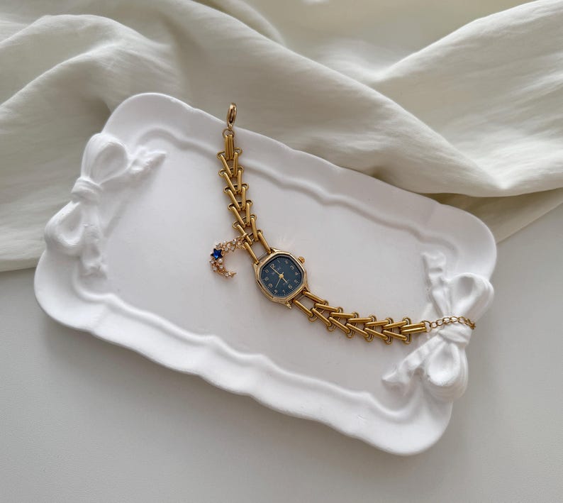 May include: A gold-toned wristwatch with a dark blue face and a decorative moon and star charm. The watch is displayed on a white, bow-shaped tray. The watch band has a unique chain design.