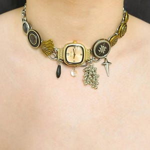 May include: A gold chain choker necklace with a variety of charms, including buttons, a watch, a cross, and a beaded tassel. The watch face is square and has a gold frame.