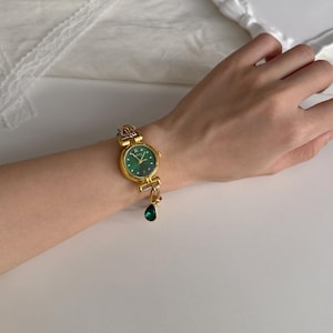 May include: A gold-toned wristwatch with a round, emerald green face and gold hands. The watch has a decorative gold band and a dangling green teardrop charm. The watch is on a wrist.