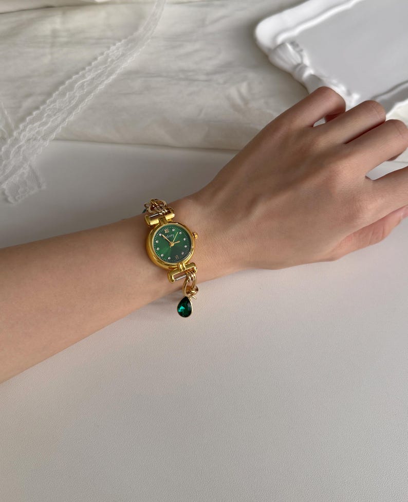 May include: A gold-toned wristwatch with a round, emerald green face and gold hands. The watch has a decorative gold band and a dangling green teardrop charm. The watch is on a wrist.