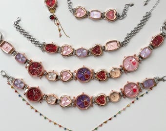 May include: A collection of heart-shaped and rectangular face watches and bracelets in shades of red, pink, and purple. The watches have silver chains and a gold-colored bezel. A heart-shaped keychain and a necklace with a crescent moon charm are also included.