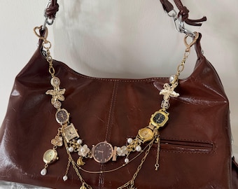 May include: A brown leather shoulder bag with a decorative gold chain featuring various watch faces, charms, and pearls. The bag has a curved shape and a long strap. The chain includes a sun, moon, and star charms.