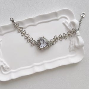May include: A silver heart-shaped watch with a white face and rhinestone accents. The watch is on a silver chain bracelet with a flower-shaped link design.