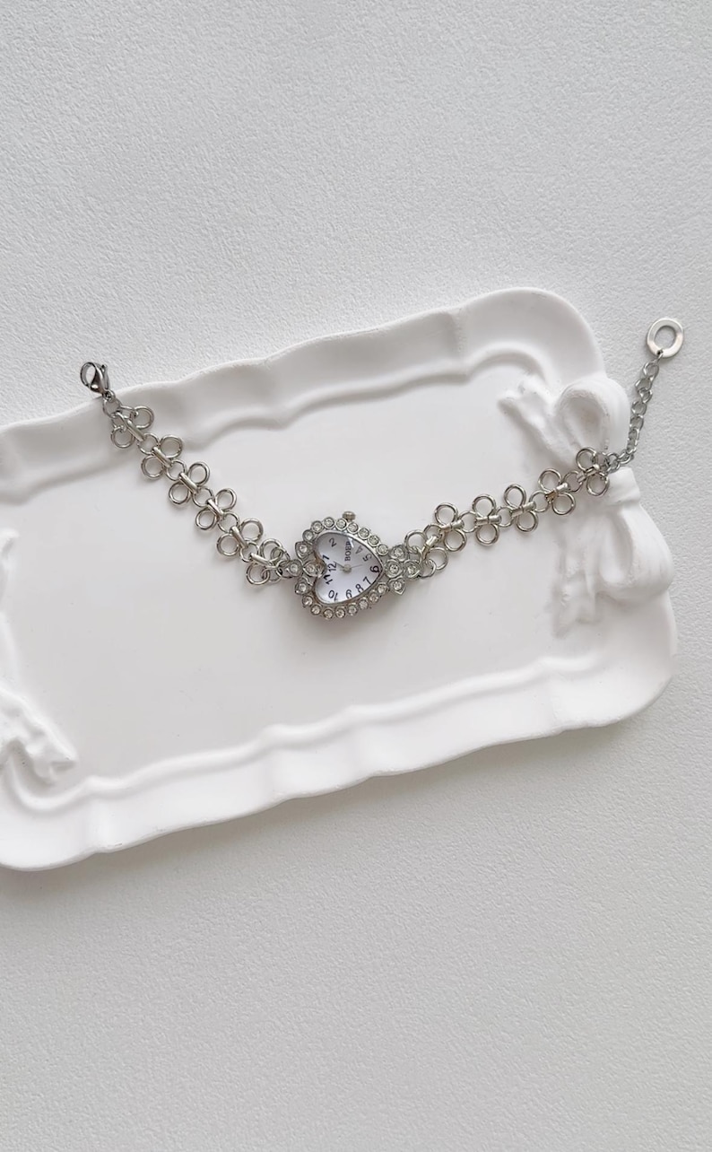 May include: A silver heart-shaped watch with a white face and rhinestone accents. The watch is on a silver chain bracelet with a flower-shaped link design.