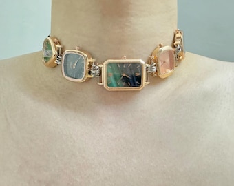 May include: A gold-tone choker necklace with five square-shaped watch faces. The watch faces have different coloured dials, including black, green, and pink.
