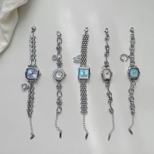 May include: Five silver-toned wristwatches with square and round faces. Each watch has a unique chain design and decorative charms. The watch faces are light blue, white, and silver. The watches are displayed on a white surface.