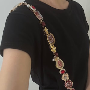 Könnte beinhalten: Ein dekorativer Schulterriemen, bestehend aus einer Reihe goldfarbener Uhrenzifferblätter, roten herzförmigen Anhängern, floralen Akzenten und kleinen weißen Perlen. Der Riemen zeigt mehrere analoge Uhrenzifferblätter mit roten Zifferblättern.