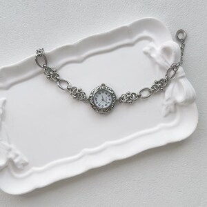 May include: A silver watch with a round face and a chain link band. The watch is on a white tray.
