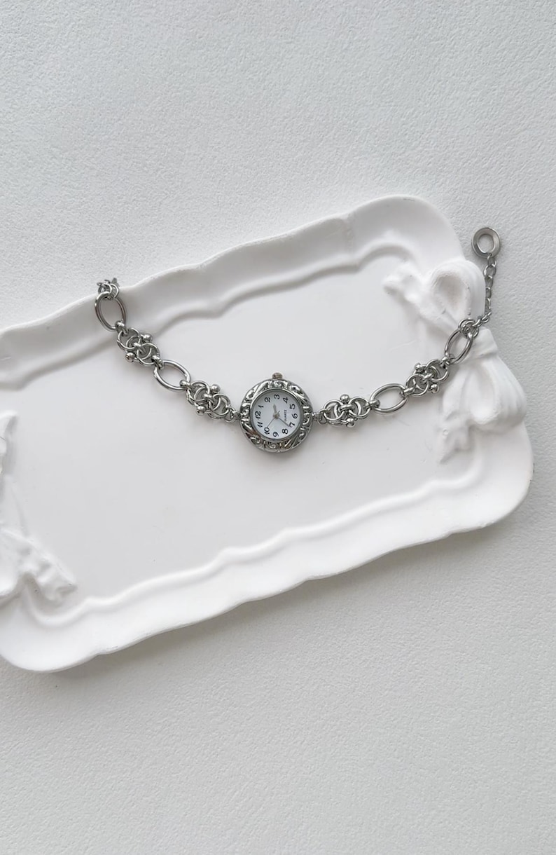 May include: A silver watch with a round face and a chain link band. The watch is on a white tray.