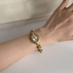 May include: Gold-toned oval wristwatch with a white face and delicate gold chain bracelet. The watch features a classic design with a textured bezel and a secure clasp. The bracelet is approximately 17.78 cm long.