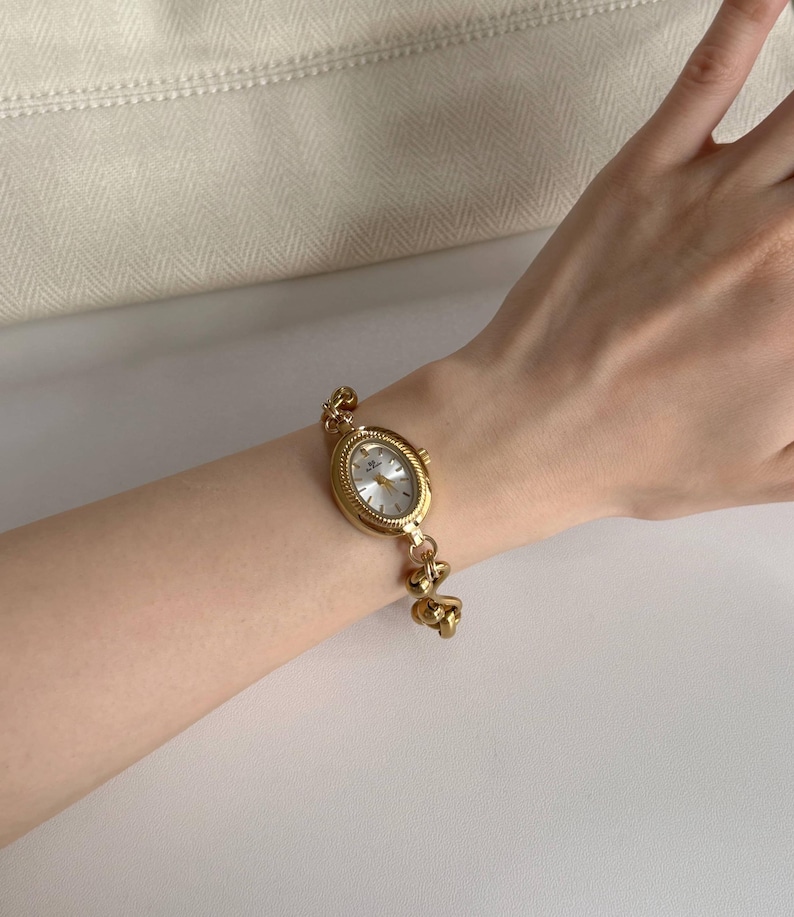 May include: Gold-toned oval wristwatch with a white face and delicate gold chain bracelet. The watch features a classic design with a textured bezel and a secure clasp. The bracelet is approximately 17.78 cm long.