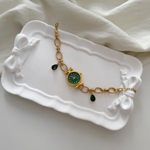 May include: Gold-toned watch bracelet with a round green watch face and dangling green teardrop-shaped charms. The watch is on a white decorative tray with bow accents. The bracelet features a chain link design.