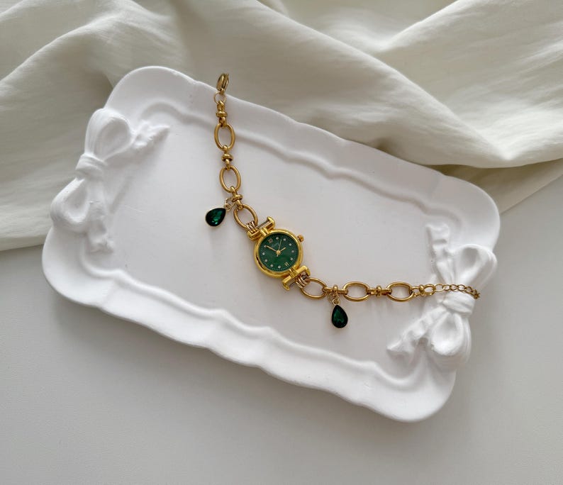 May include: Gold-toned watch bracelet with a round green watch face and dangling green teardrop-shaped charms. The watch is on a white decorative tray with bow accents. The bracelet features a chain link design.