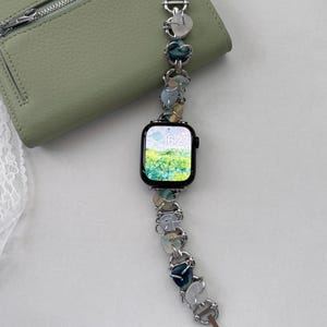 Peut inclure: Une montre intelligente noire avec un bracelet décoratif orné de perles en forme de cœur et rondes dans des tons de bleu, vert et blanc. La montre est présentée sur un portefeuille en cuir vert clair.