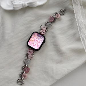 Puede incluir: Un reloj inteligente negro con una correa decorativa. La correa es plateada con dijes de corazón y flores rosas, y un pequeño dije de mariposa. La esfera del reloj muestra un gráfico rosa. La hora es 16:15.