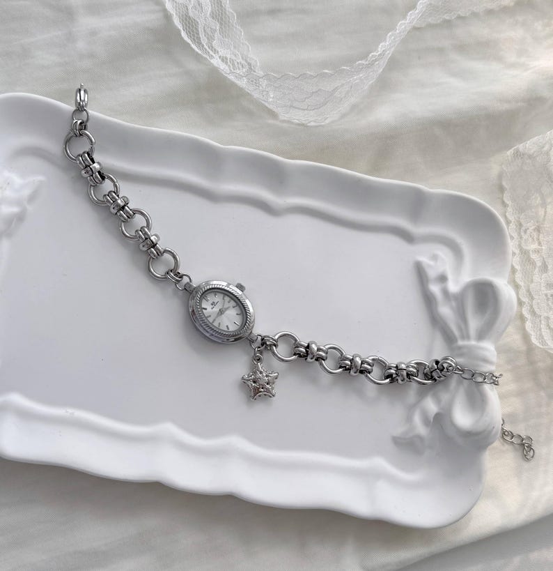 May include: A silver bracelet watch with an oval face and a star charm. The watch has a chain-link design and a white watch face. The star charm is embellished with small, clear stones. The watch is displayed on a white, decorative tray.