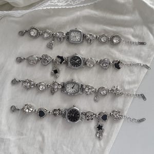 Może przedstawiać: Cztery srebrne zegarki z zawieszkami o różnych kształtach tarczy i dekoracyjnych zdobieniach. Każdy zegarek ma unikalny wzór tarczy i jest ozdobiony zawieszkami, w tym sercami, kwiatami i gwiazdami. Zegarki są prezentowane na białej powierzchni.