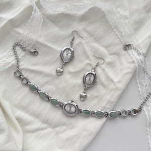 May include: A silver-toned jewelry set featuring a watch-style choker necklace and matching earrings. The necklace has oval-shaped green stones and watch faces. The earrings have watch faces with heart-shaped charms. The set is displayed on a white fabric.