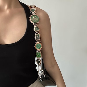May include: A gold chain strap with multiple green and gold watches attached. The strap is worn over a black tank top and beige pants.