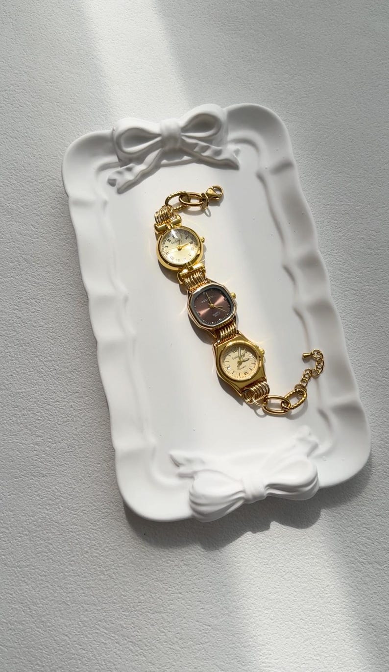 May include: A gold-toned watch bracelet with three different watch faces. The bracelet is on a white ceramic tray with a bow design.