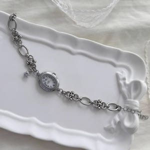 May include: A silver bracelet watch with an oval face and a white dial. The watch is attached to a chain with decorative links and a small charm. The watch is presented on a white tray.