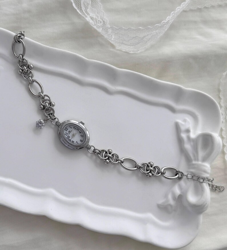 May include: A silver bracelet watch with an oval face and a white dial. The watch is attached to a chain with decorative links and a small charm. The watch is presented on a white tray.