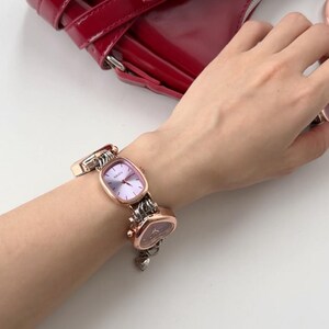May include: A rose gold watch with a square face and a heart-shaped charm on the band. The watch is on a person's wrist.