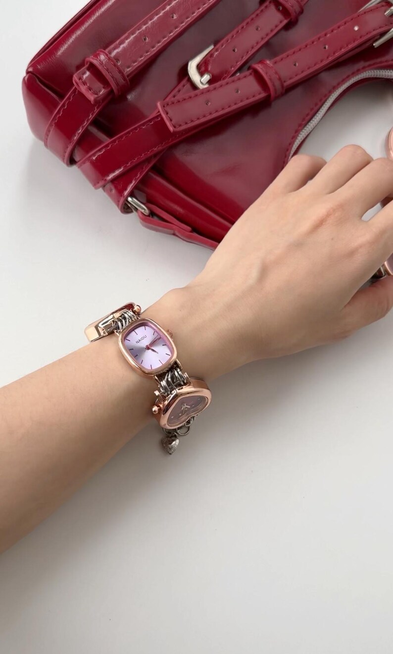 May include: A rose gold watch with a square face and a heart-shaped charm on the band. The watch is on a person's wrist.