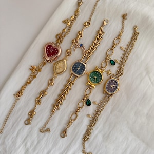 May include: Five gold-toned wristwatches with various face shapes and colors, including red heart, oval, and round. Each watch has a unique chain design with decorative charms. The watches are displayed on a white, textured surface.