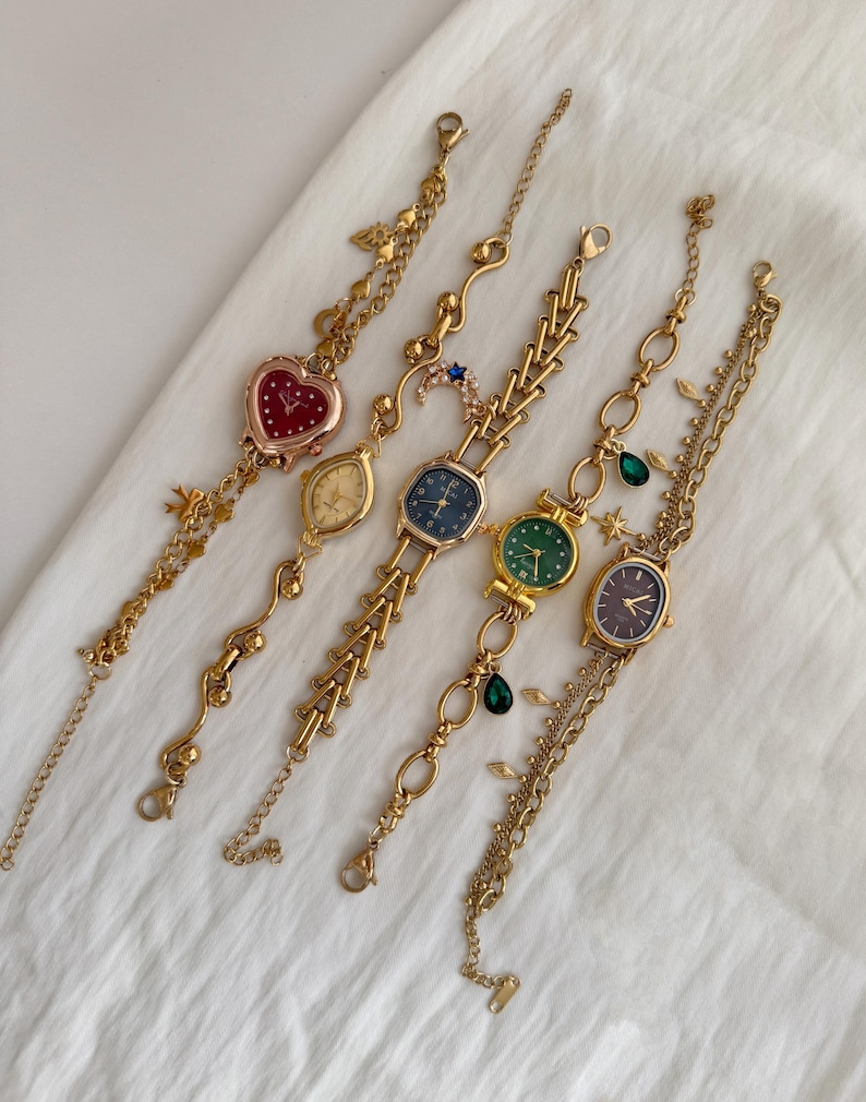 May include: Five gold-toned wristwatches with various face shapes and colors, including red heart, oval, and round. Each watch has a unique chain design with decorative charms. The watches are displayed on a white, textured surface.