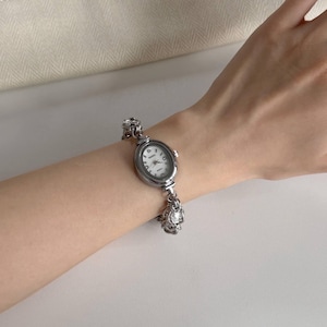 May include: Silver oval wristwatch with a white face and black numerals. The watch has a decorative chain-link band. The watch is worn on a wrist.