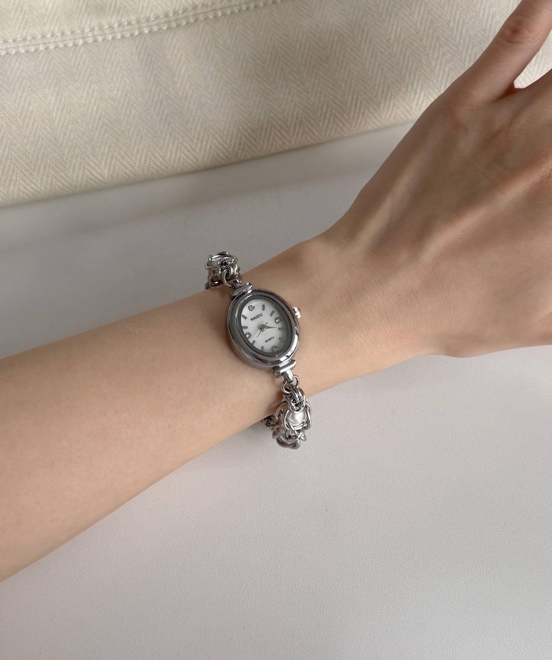 May include: Silver oval wristwatch with a white face and black numerals. The watch has a decorative chain-link band. The watch is worn on a wrist.