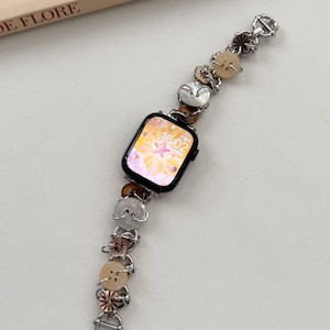 May include: A black smartwatch with a decorative strap. The strap is adorned with beige, brown, and iridescent white buttons, linked by silver-toned metal. The watch face shows a floral design. The watch strap is approximately 18 cm long.