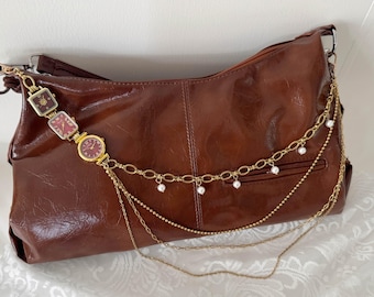 May include: A brown leather shoulder bag with gold chain and pearl detailing. The bag features a decorative chain of three vintage watches. The watches have red faces and gold frames. The bag has a zip closure and a pocket.