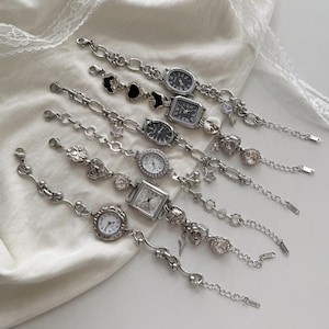 May include: An assortment of silver chain watches with different face designs, such as oval, square, and round. Some watches have decorative charms like hearts, stars, and floral details. The watches are presented on a white, textured background.