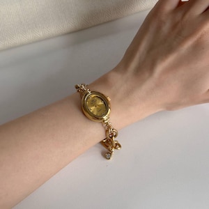 May include: A gold wristwatch with an oval face and a matching bracelet. The watch has a gold dial and a small charm with a clear gemstone. The bracelet is connected with decorative links.