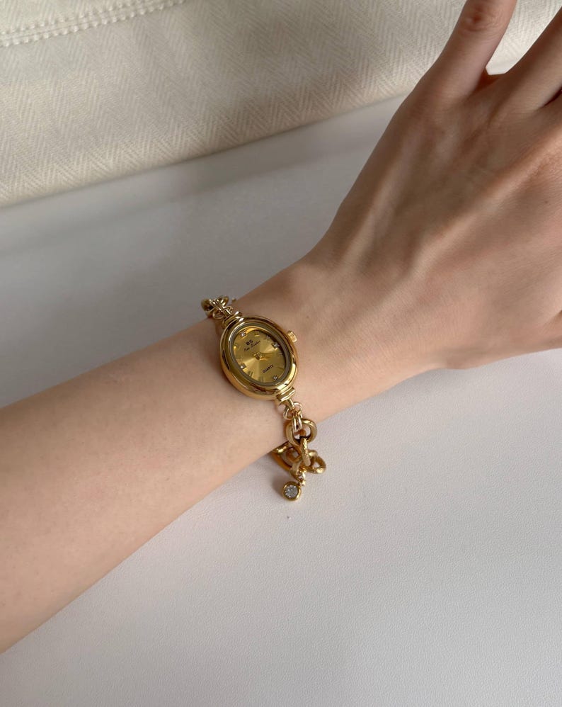 May include: A gold wristwatch with an oval face and a matching bracelet. The watch has a gold dial and a small charm with a clear gemstone. The bracelet is connected with decorative links.