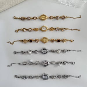 May include: Six vintage-style wristwatches with ornate bands. Three are gold-toned with oval or round watch faces and decorative links. The other three are silver-toned, featuring round watch faces and embellished with pearls and crystals. The watches are displayed on a white surface.