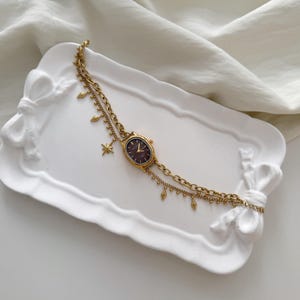 May include: A gold-toned wristwatch with a dark brown face and a delicate chain. The watch features a star charm and small dangling accents. It rests on a white, bow-shaped tray, set against a white fabric backdrop.