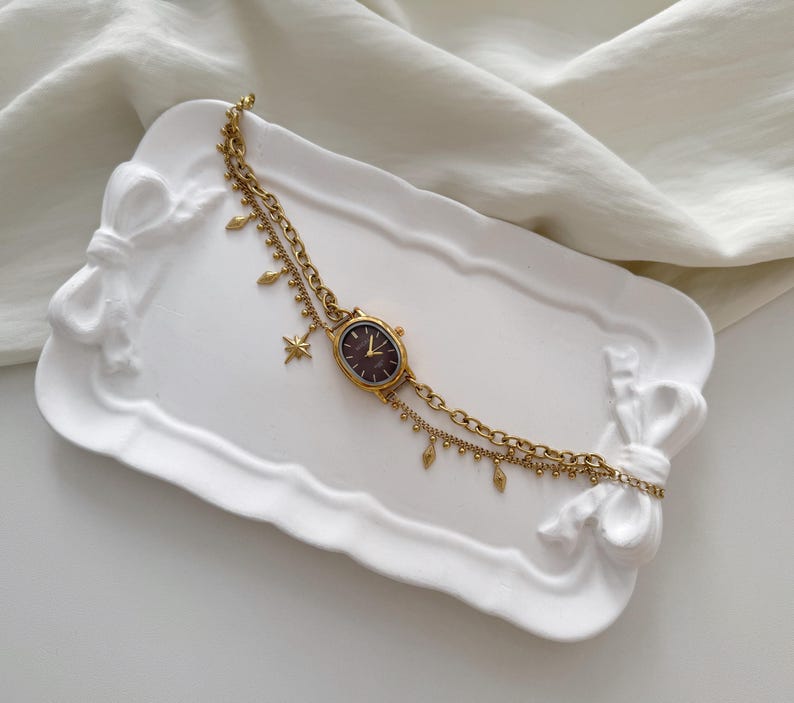 May include: A gold-toned wristwatch with a dark brown face and a delicate chain. The watch features a star charm and small dangling accents. It rests on a white, bow-shaped tray, set against a white fabric backdrop.