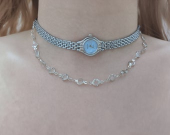 May include: A silver watch with a blue face on a silver chain necklace. The necklace has clear crystal beads.
